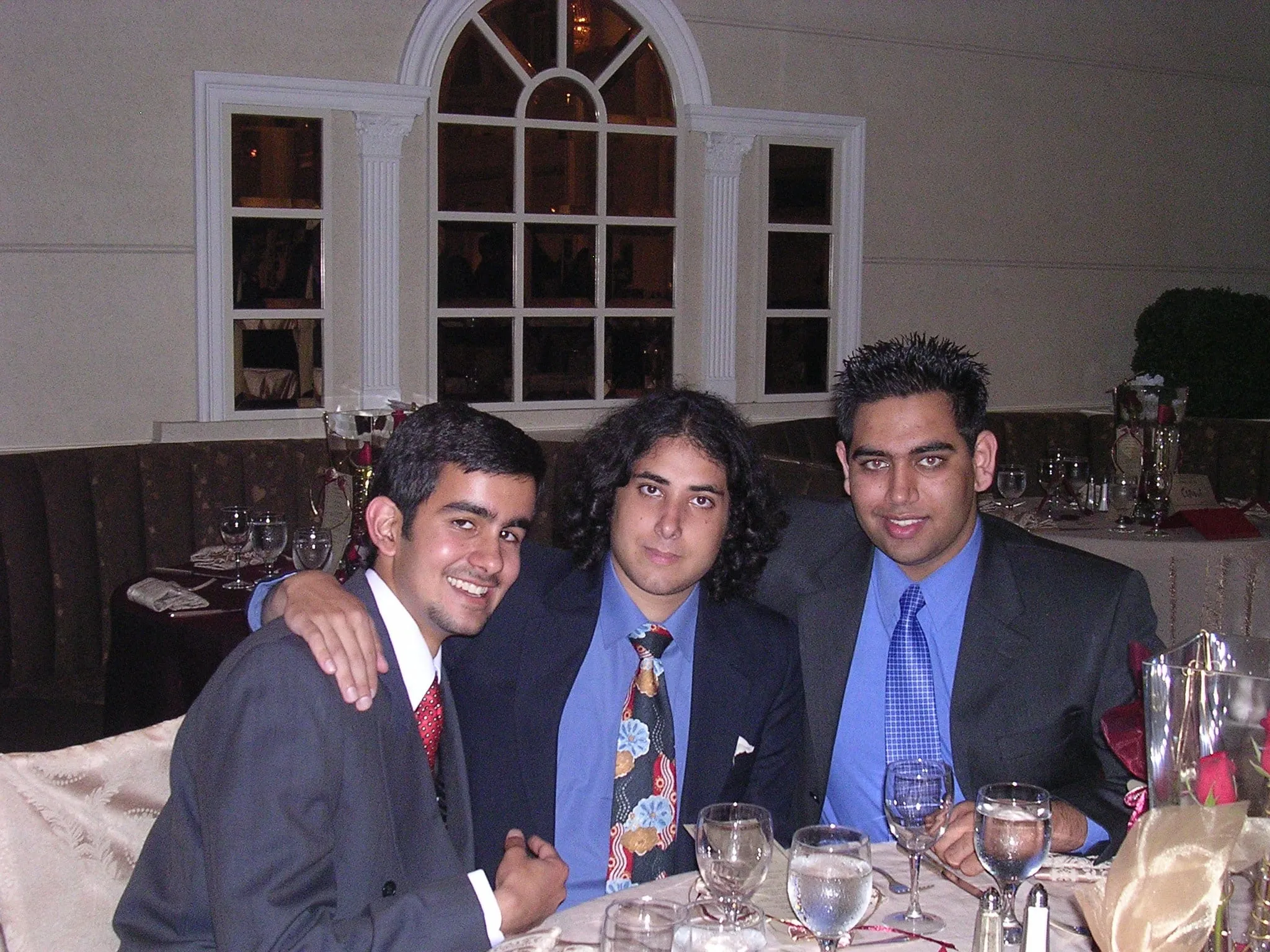 Three young men in suits at a dinner table, arms around each other