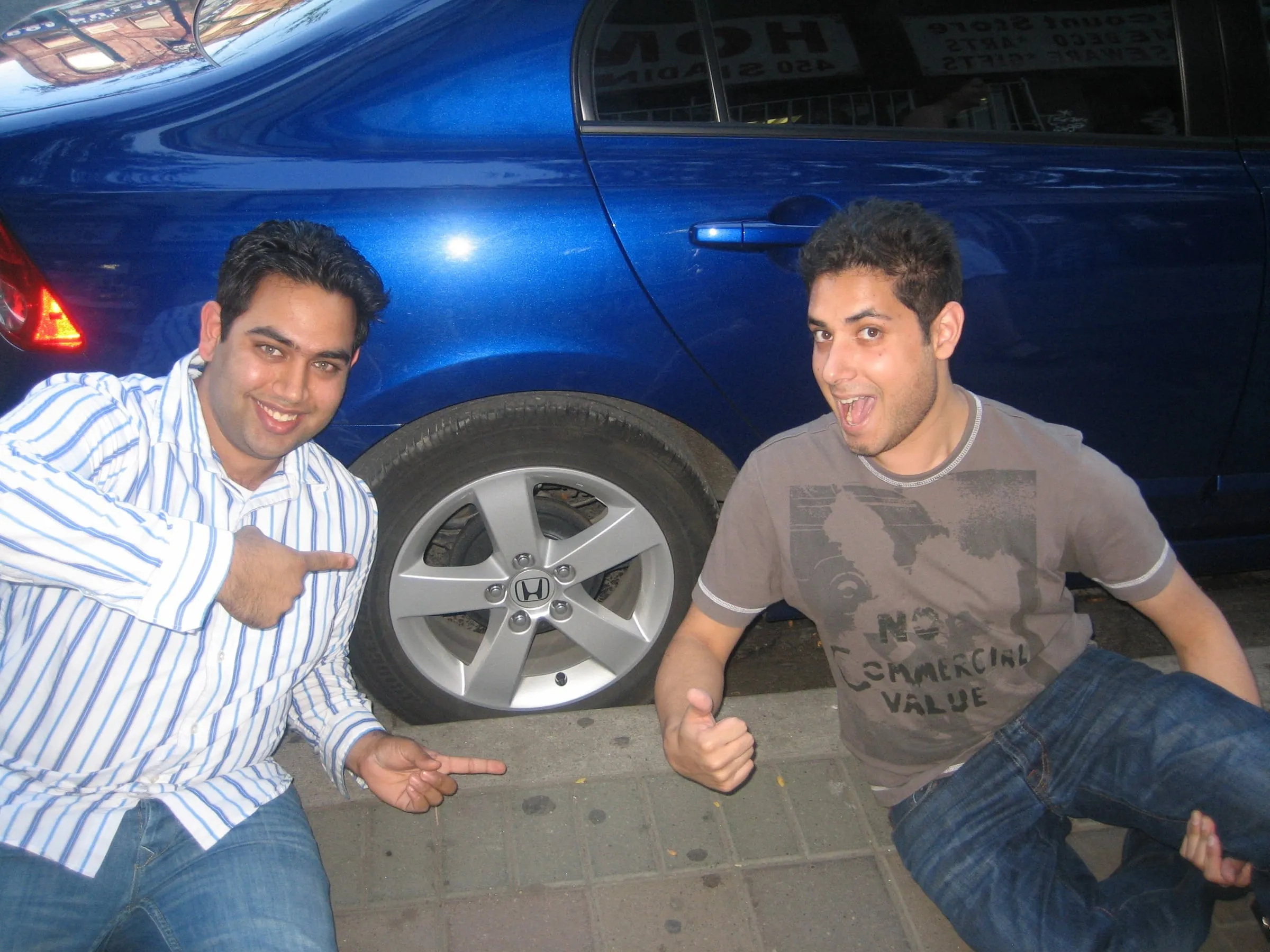 Two young men crouched beside a blue car, pointing at the camera