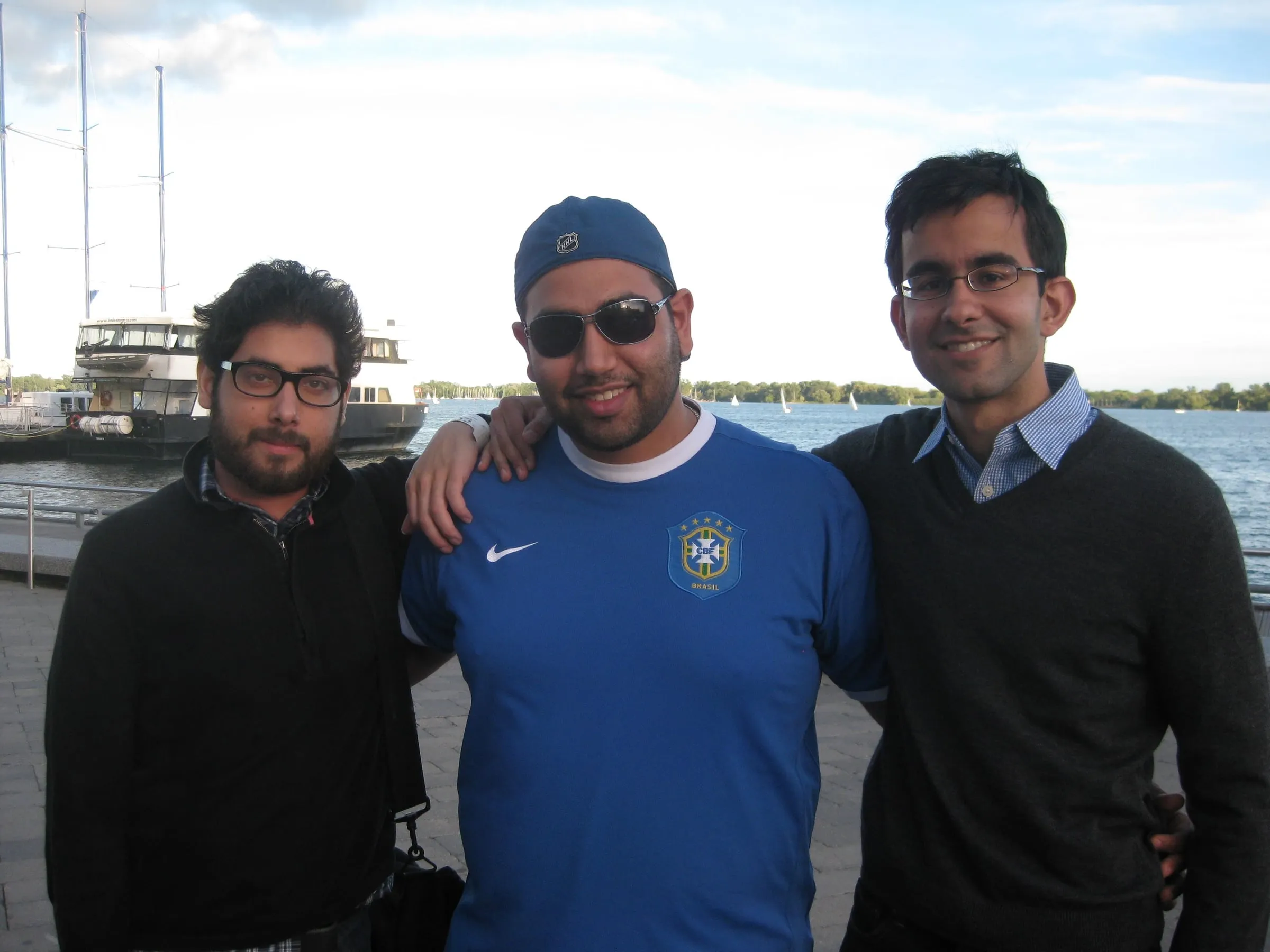 Three friends standing together by the water, boats behind them