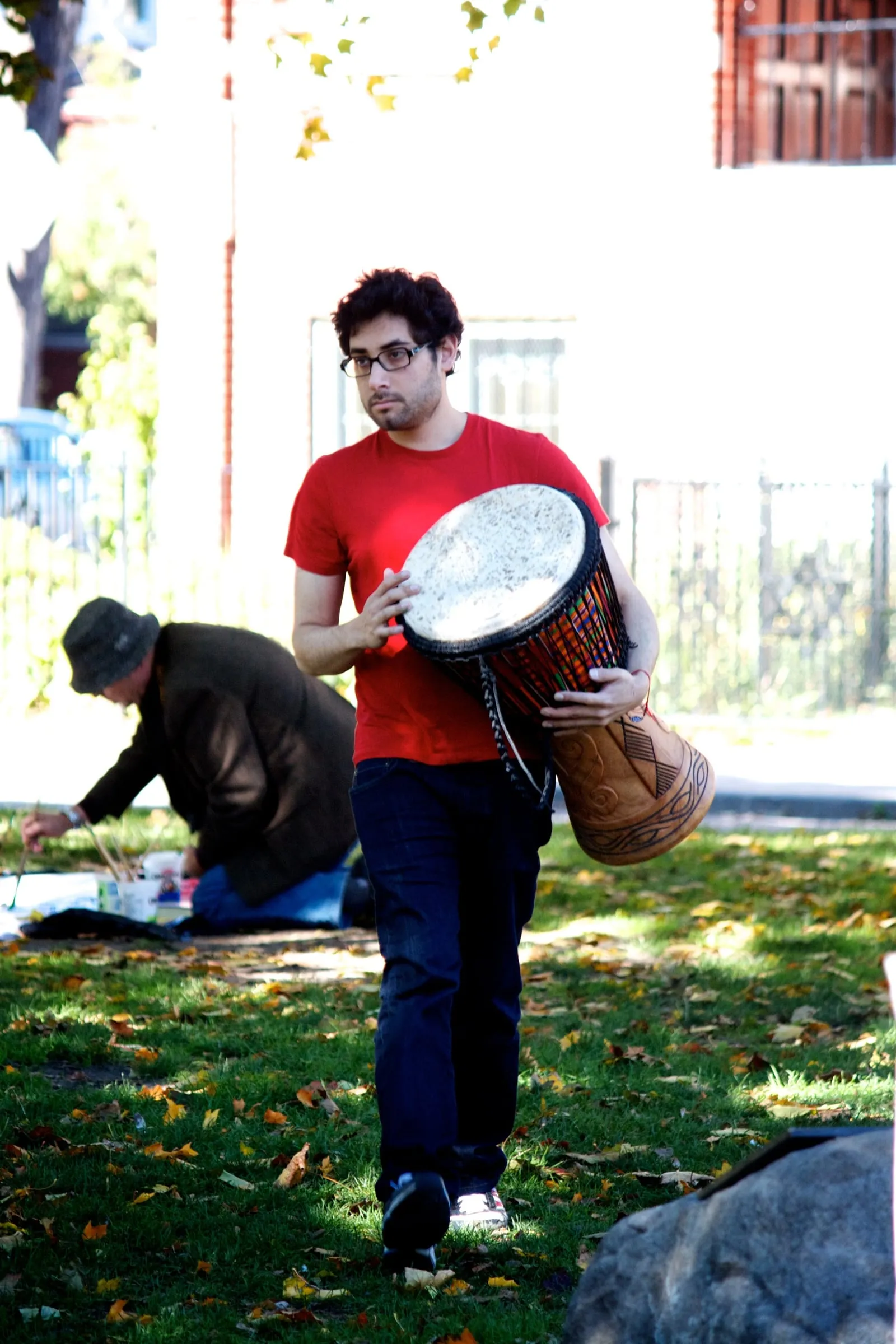 A young man in a red T-shirt and glasses carrying a hand drum across a park