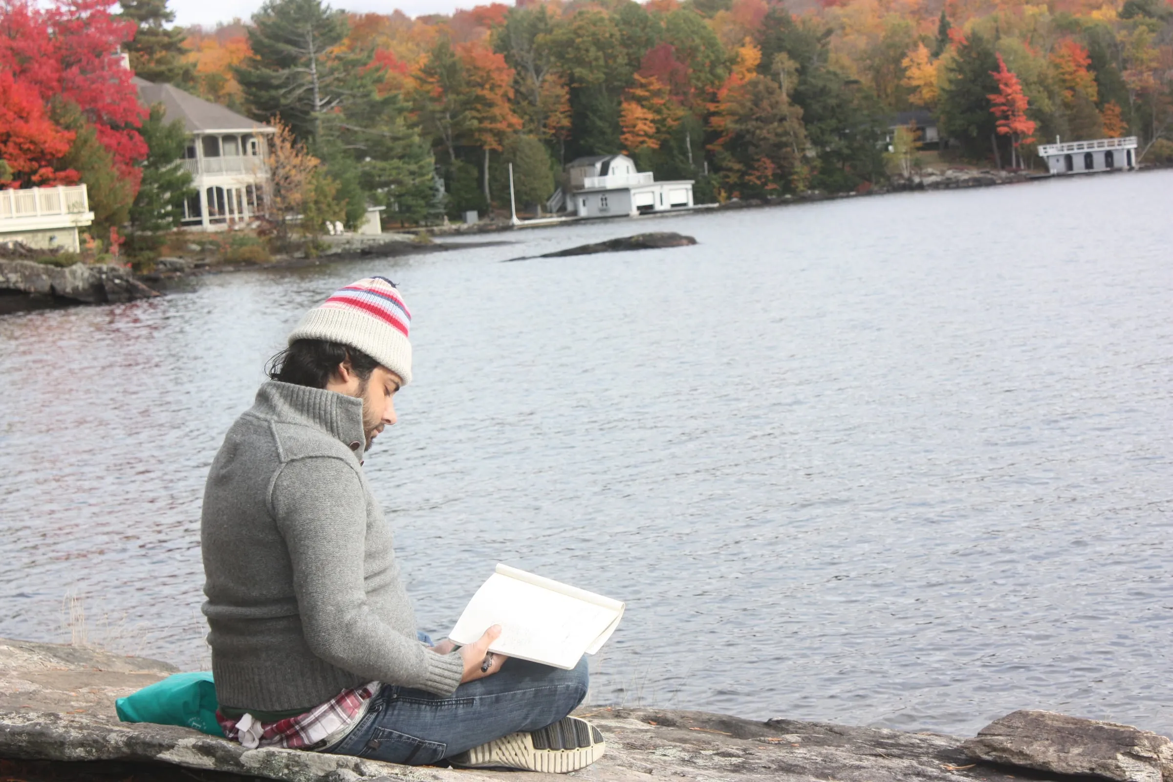 A person in a knit hat reading on the rocks by a lake, autumn trees on the far shore